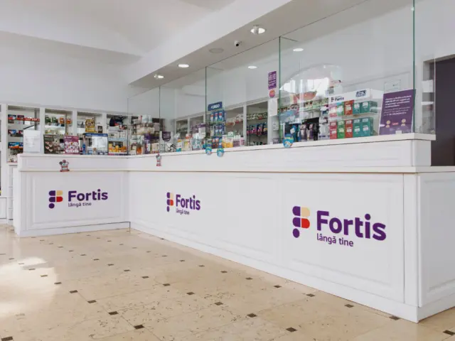 Trei sigle Fortis cu litere volumetrice mov, montate pe tejgheaua albă din interiorul farmaciei.