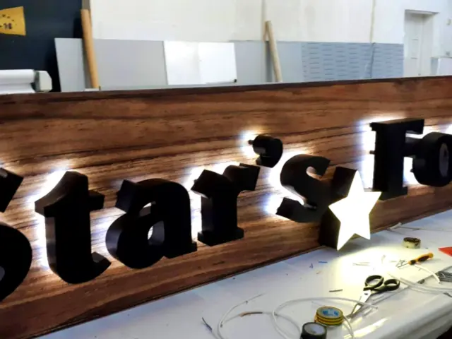 Litere volumetrice negre Star’s Food, iluminate din spate pe un panou cu aspect de lemn, în atelier.