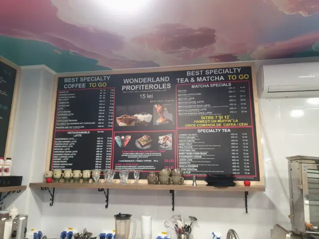 Panou meniu montat deasupra tejghelei, cu sortimente de cafea, matcha, ceai și deserturi.