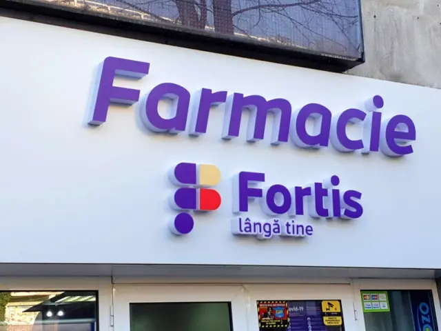 Firmă Farmacie Fortis cu litere volumetrice mov și logo colorat, montată pe panoul alb deasupra intrării.