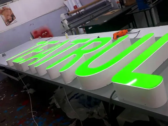 Litere volumetrice cu fețe verzi luminoase, așezate pe masa de lucru din atelier.