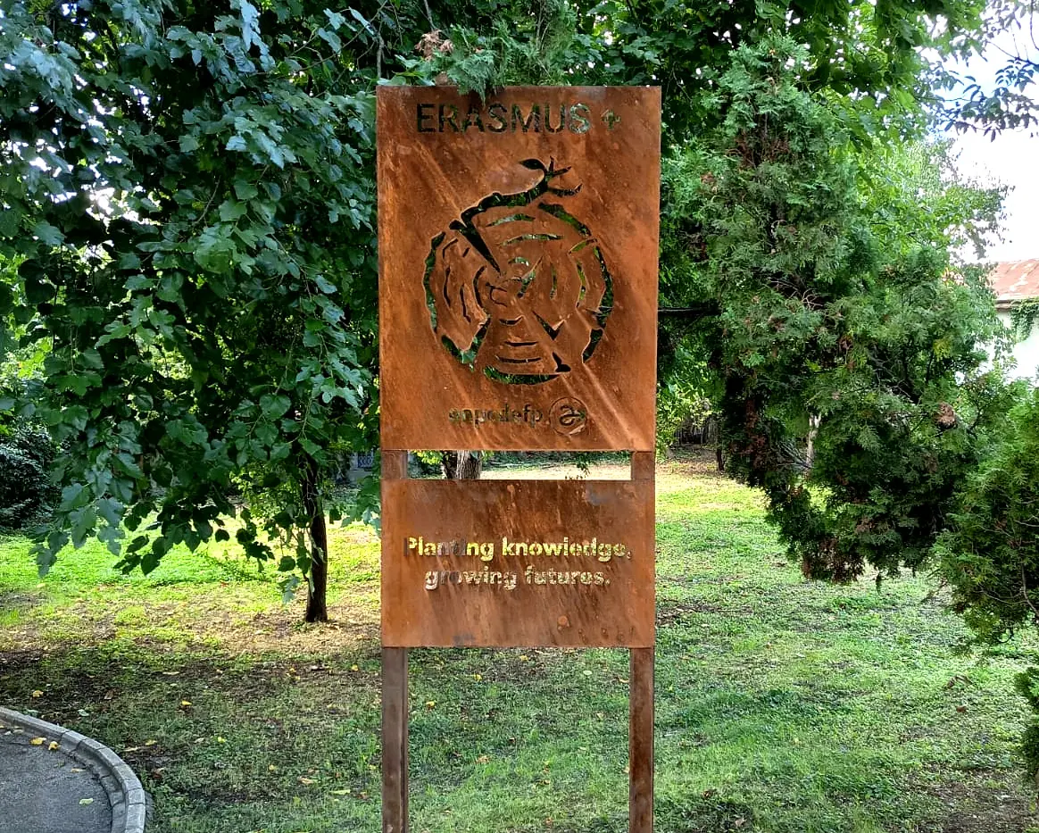 Panou metalic Erasmus+ cu text și decupaje decorative, montat pe doi stâlpi într-o zonă verde.