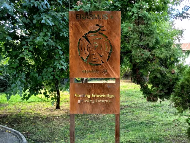Panou metalic Erasmus+ cu text și decupaje decorative, montat pe doi stâlpi într-o zonă verde.