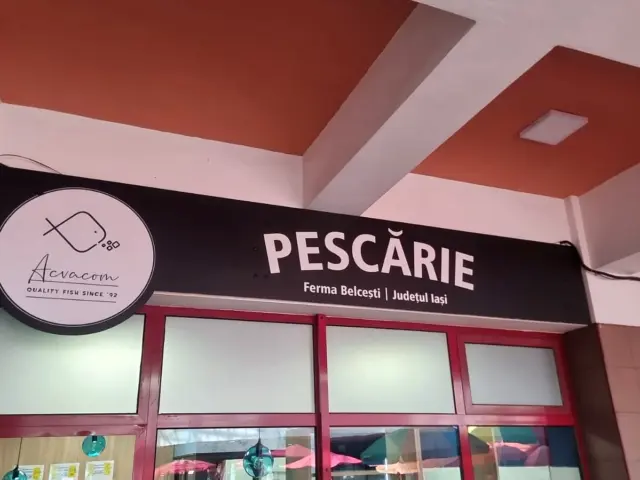 Panou negru Acvacom montat deasupra vitrinei pescăriei, cu siglă rotundă și text alb.