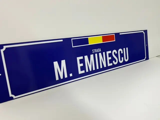 Semn stradal albastru inscripționat „Strada M. Eminescu”, cu tricolor și chenar alb decorativ.