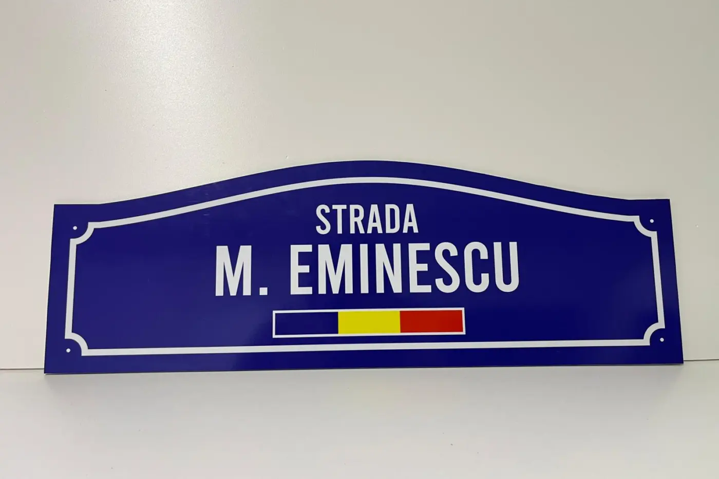 Plăcuță stradală albastră cu contur alb, inscripția „Strada M. Eminescu” și tricolorul României.