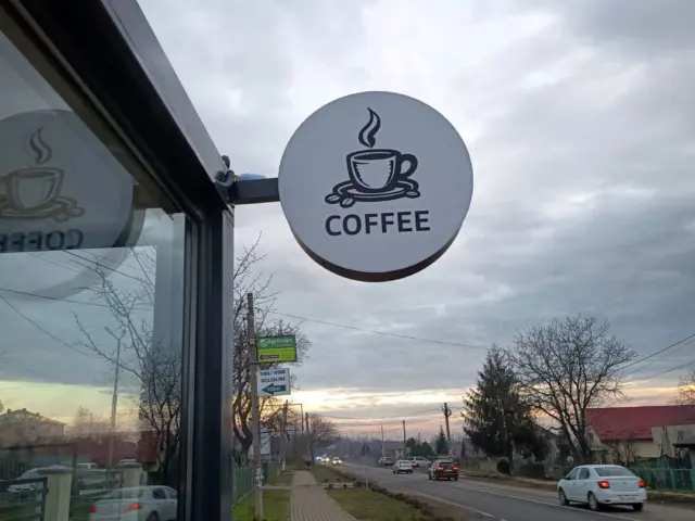 Firmă luminoasă rotundă, montată perpendicular pe fațadă, cu ceașcă stilizată și textul COFFEE.