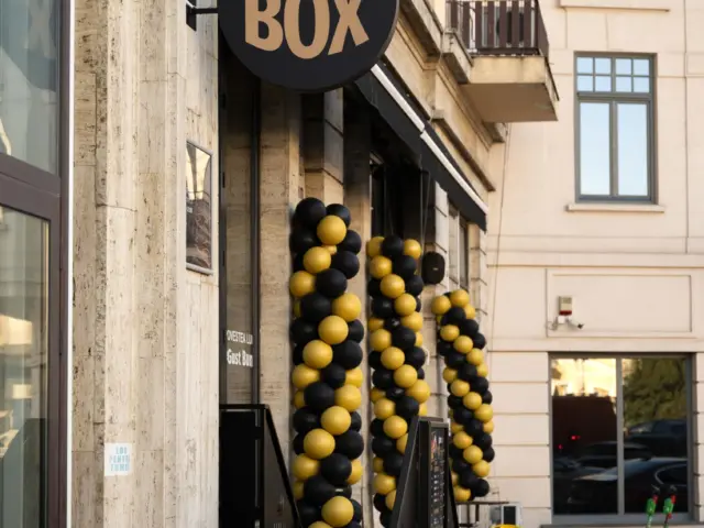 Firmă rotundă The BOX montată perpendicular deasupra intrării, lângă arcade din baloane negre și galbene.