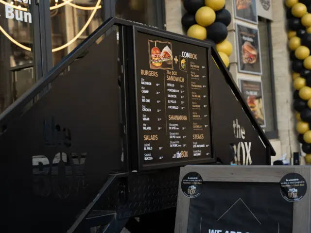 Panou de meniu the BOX montat lângă scările restaurantului, cu preparate și prețuri afișate.