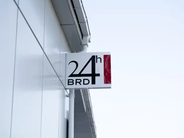 Firmă luminoasă BRD 24h, montată perpendicular pe fațada albă a clădirii.
