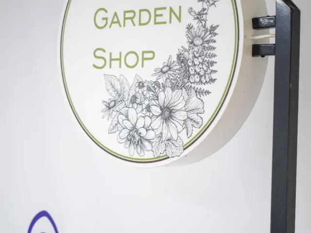 Firmă rotundă The Garden Shop, montată perpendicular pe suport, cu text verde și ilustrații botanice.