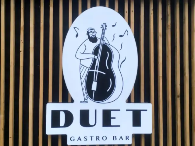 Firmă luminoasă DUET Gastro Bar, cu ilustrația unui contrabasist, montată pe un perete cu lamele verticale.