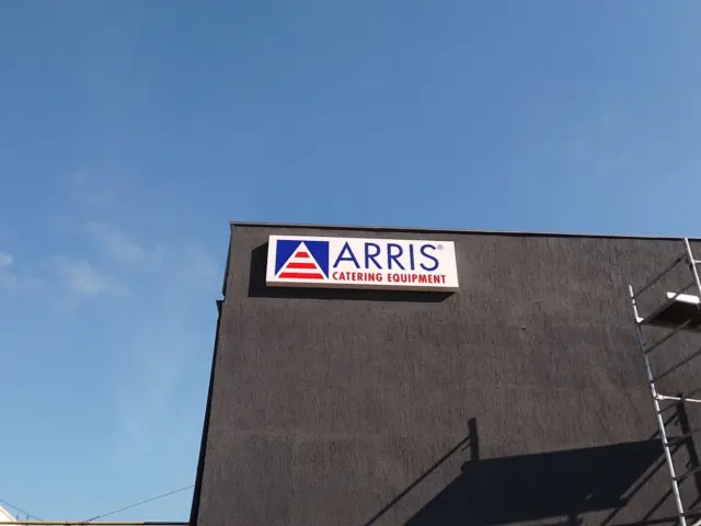 Casetă ARRIS Catering Equipment montată sus pe fațada închisă la culoare, lângă o schelă.