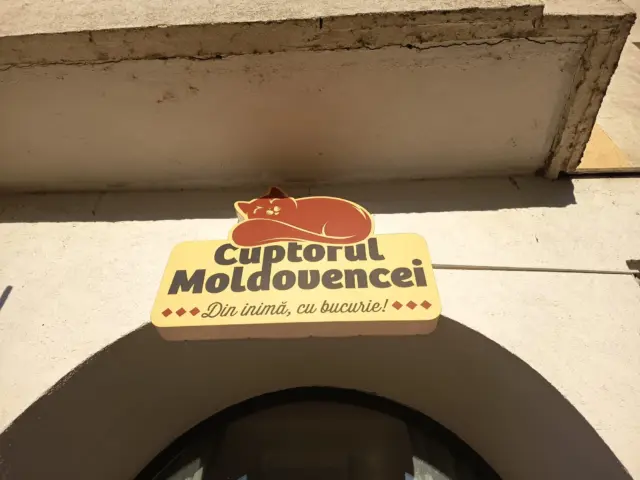 Firmă Cuptorul Moldovencei, cu pisică ilustrată și slogan, montată deasupra unei intrări arcuite.