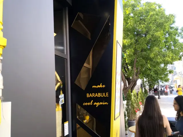 Panou negru Cartuf cu inserții geometrice oglindă și text galben, montat lângă intrarea stradală.