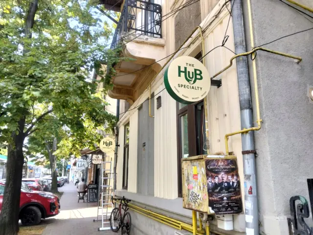 Două semne rotunde The Hup Specialty, montate perpendicular pe fațadă, de-a lungul unui trotuar cu arbori.
