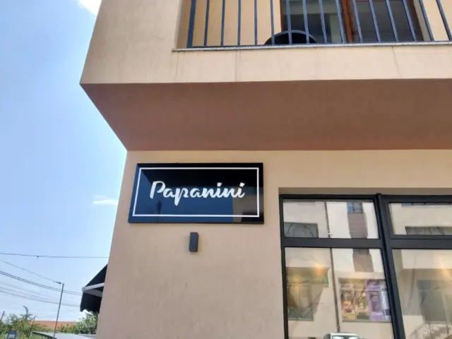 Firmă rectangulară Papanini, cu fundal negru și contur alb, montată lângă vitrina clădirii.