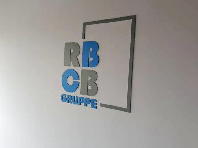 Logo-ul volumetric RCBB Gruppe, în albastru și gri, montat pe un perete alb de interior.