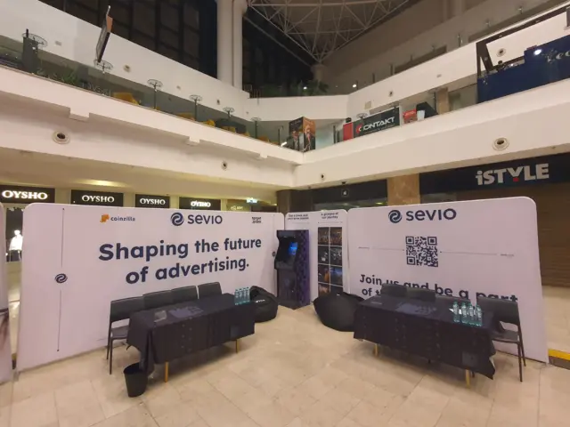 Stand Sevio cu pereți textili curbați, mese și scaune, amenajat într-un spațiu comercial.
